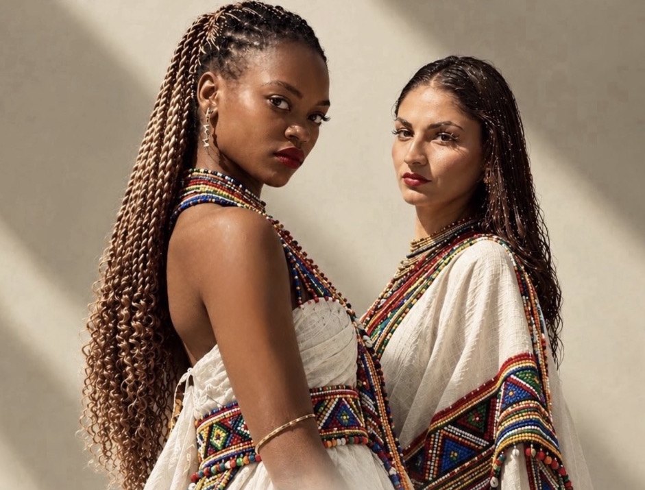 Two women in beaded, embroidered white dress against a sunlit wall, an Omniguild Heritage Day cover